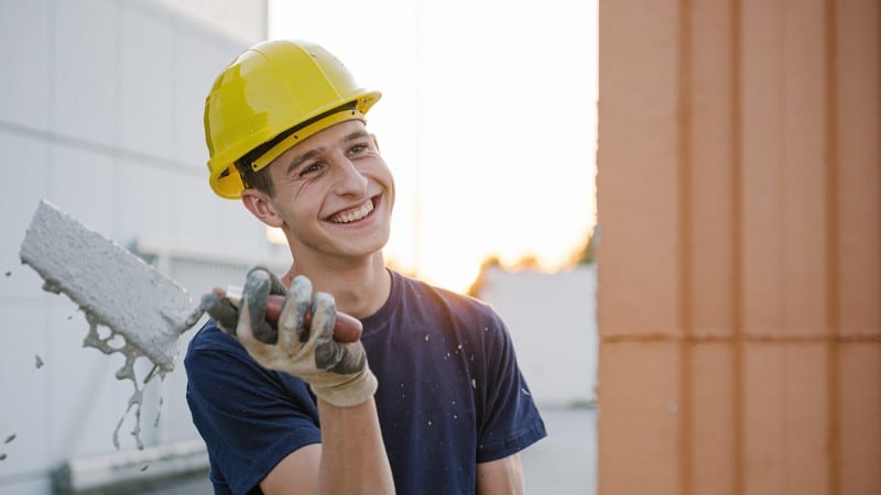 Junger lachender Mann mit Bauhelm bei Verputzarbeiten auf der Baustelle