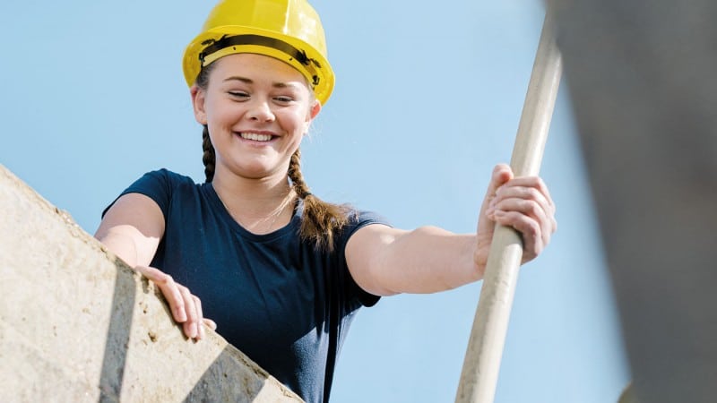 Junge, lachende Frau mit Bauhelm, die von einem Bauwerk herunterblickt